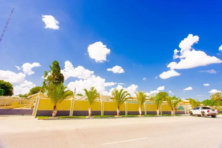 View of parking area and facade of Cubita Guesthouse in Oshakati with palm trees and clear sky.